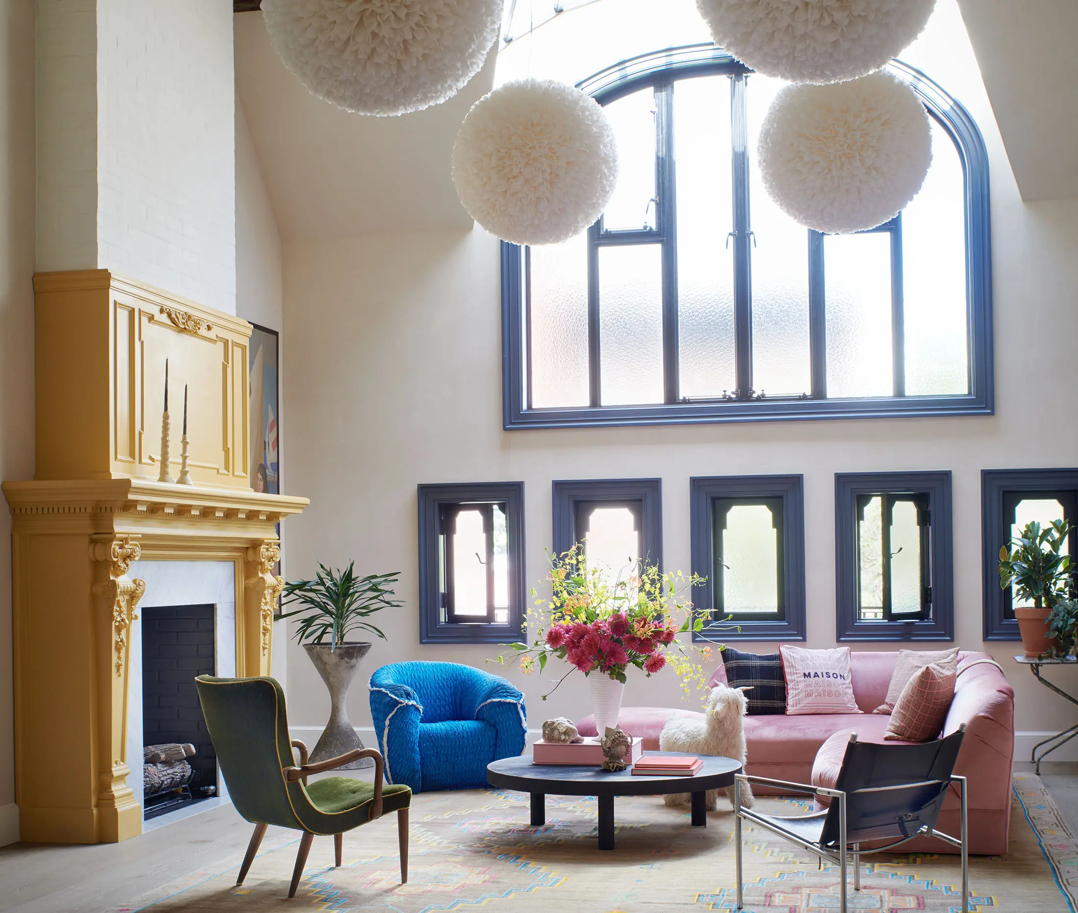 A stylish living room with a high, arched window. Sunlight streams through, illuminating a pink sofa, a blue chair, and a green armchair around a round coffee table with floral arrangements. A mustard-yellow fireplace on the left wall adds warmth. Large white pendant lights hang from the ceiling.
