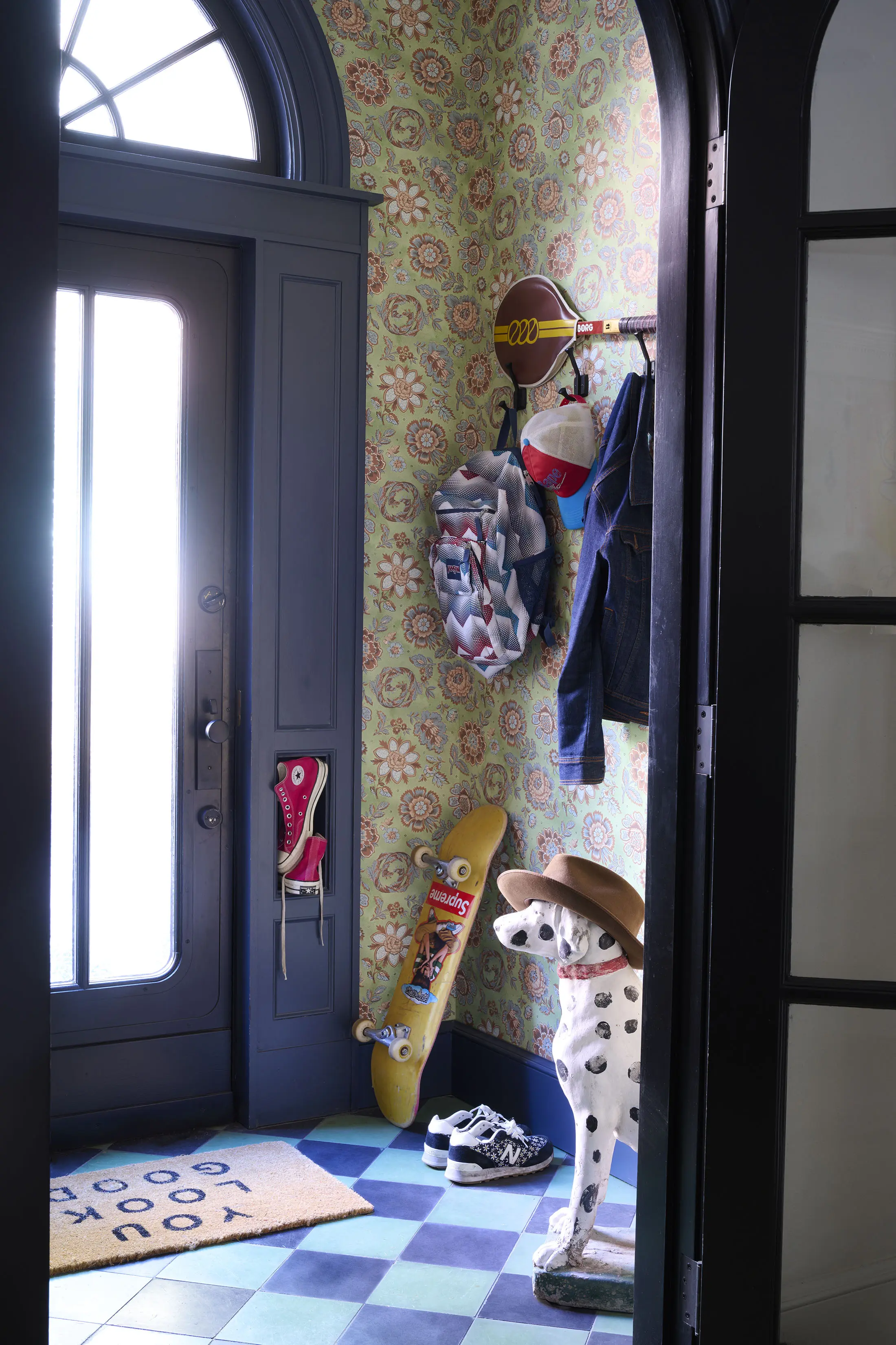 Colorful entryway with floral wallpaper and a blue arched door. A wall hook holds hats and a denim jacket, with a skateboard propped against the wall. A Dalmatian statue with a hat stands on a blue and green tiled floor next to sneakers and a YOU LOOK GOOD doormat.