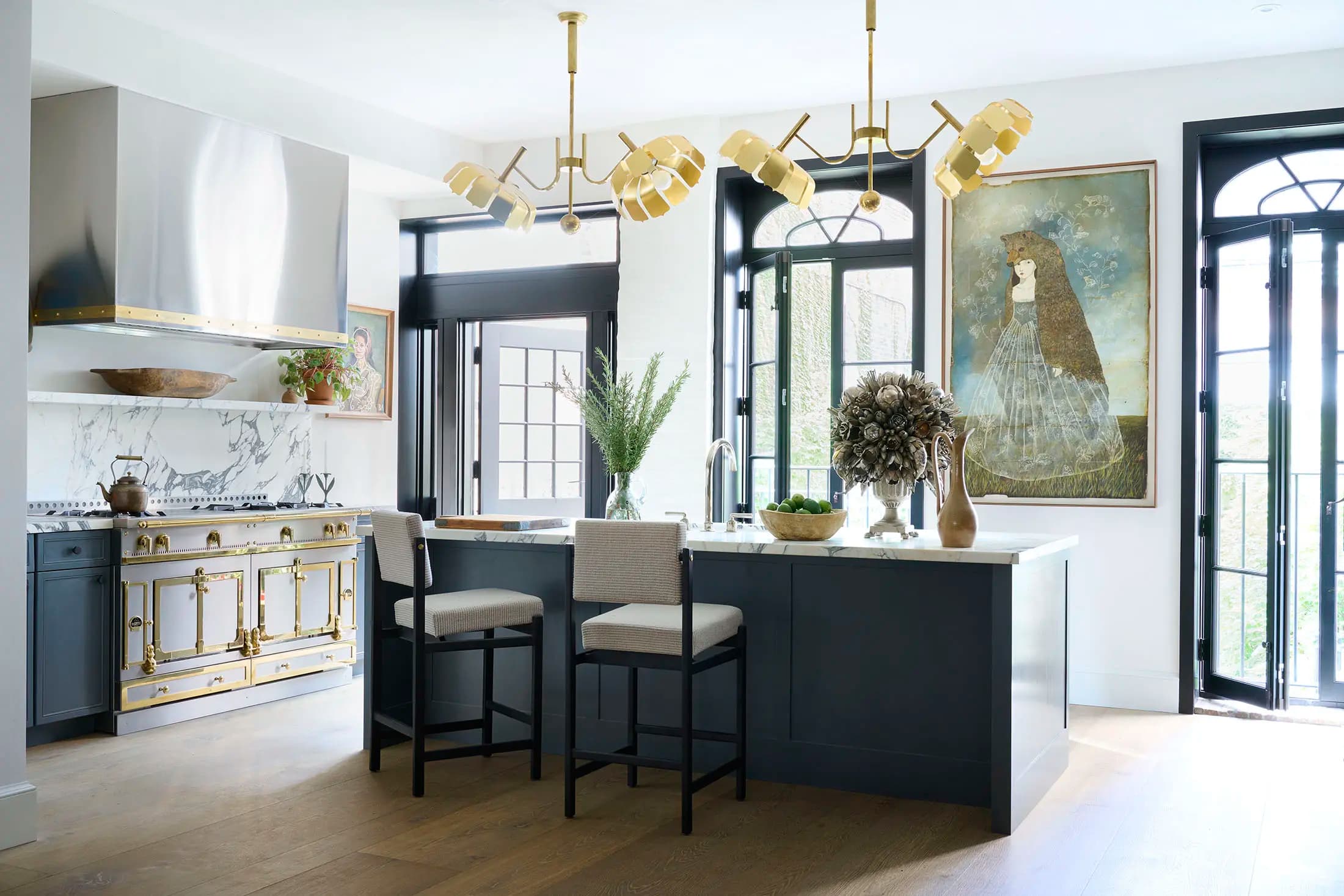 A modern kitchen with a gold-trimmed stove, dark cabinetry, and a marble backsplash. Two chairs are at an island with a green vase and brown bowl. A large painting hangs on the wall. Two geometric pendant lights hang from the ceiling. Large windows bring in natural light.
