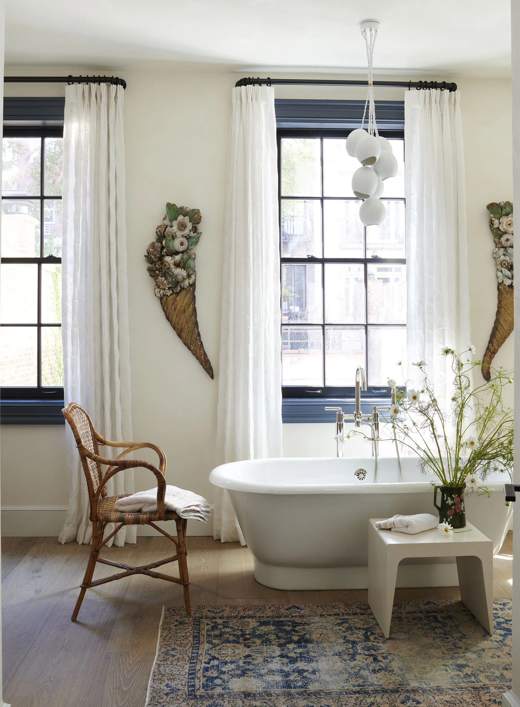 A bathroom with a freestanding white tub, a modern stool with a towel and flowers, and a wicker chair. Two tall windows with sheer white curtains, flanked by floral wall decor, allow natural light. A vintage-style rug covers the wooden floor, and an artistic white light fixture hangs above.