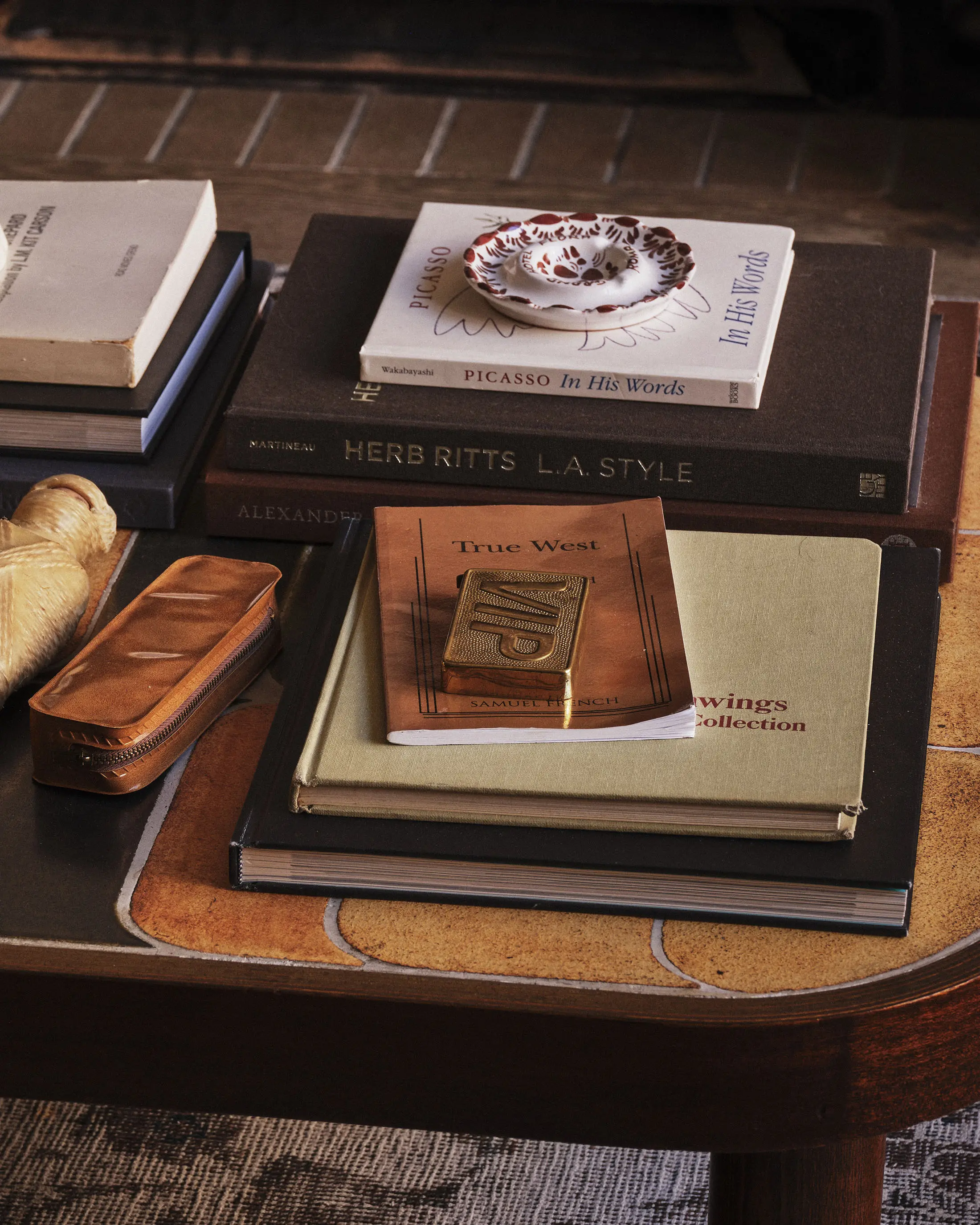 A coffee table with stacks of books, including Herb Ritts: L.A. Style, Picasso In His Words, and Alexander McQueen Story. A small dish holds seashells. A decorative box and a zippered leather case are also on the table, which features an inlaid design on its surface.
