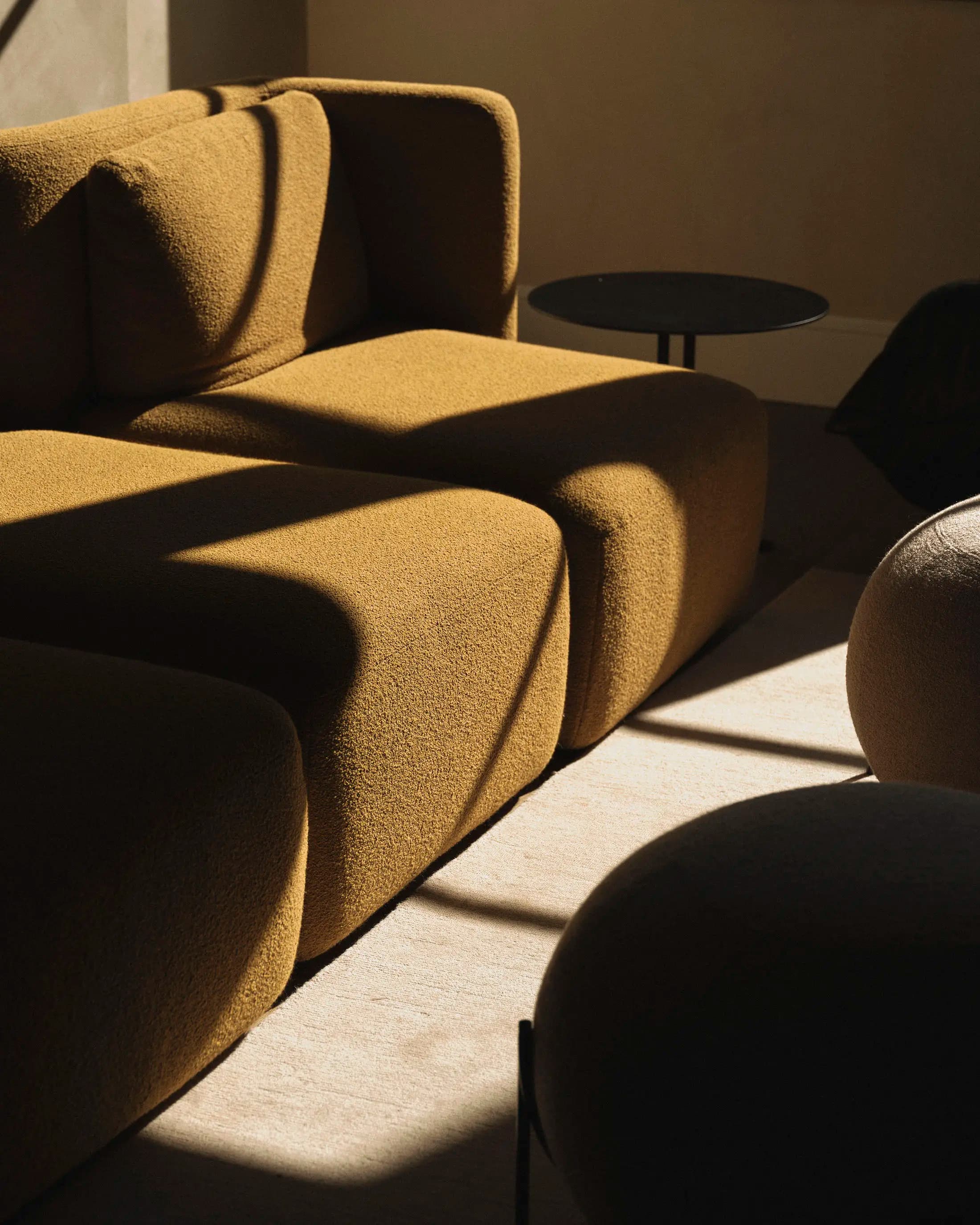A cozy living room scene featuring a mustard-colored, textured sofa with pronounced cushions. Sunlight casts sharp, contrasting shadows across the sofa and floor. A small, round black side table is partly visible in the background, adding to the modern and minimalist decor.