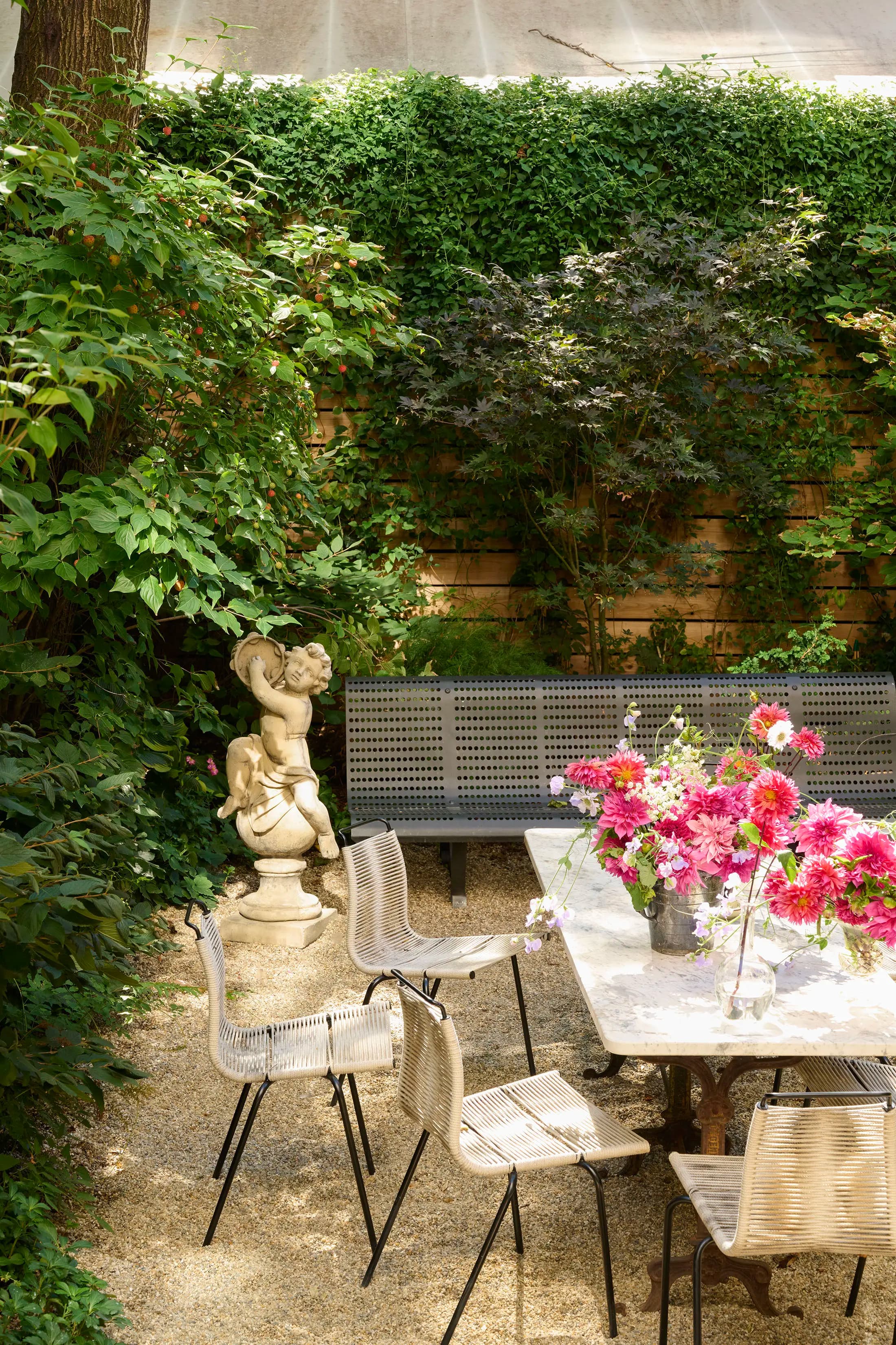 Outdoor garden setting with a long table featuring a colorful flower arrangement. Surrounding the table are wicker chairs on a gravel surface. A gray bench sits against a lush green ivy-covered wall. A classical stone sculpture of a cherub is placed nearby.