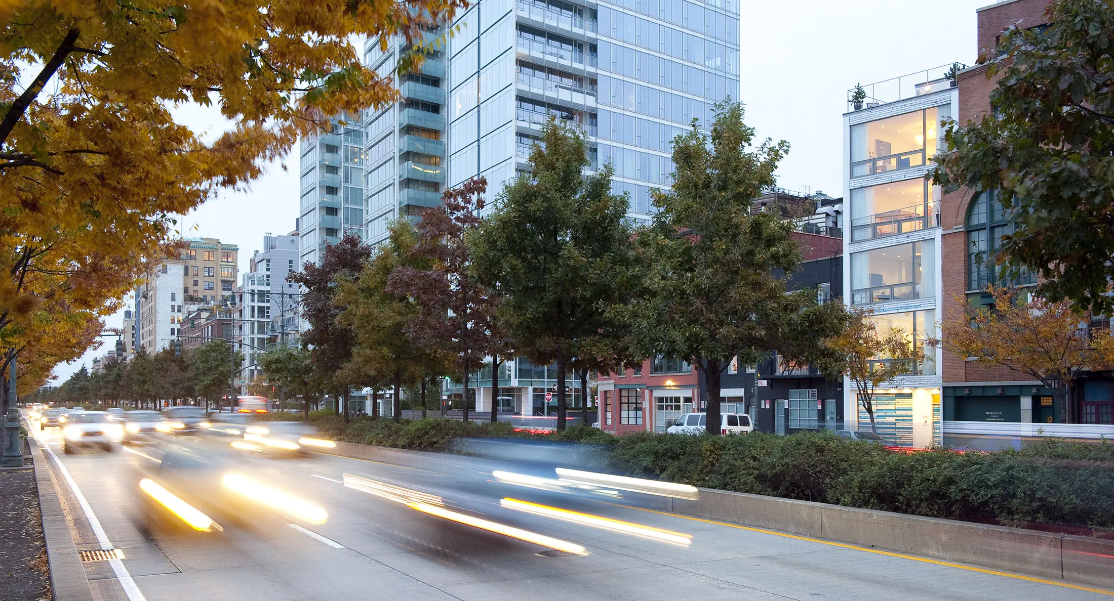 A bustling urban street scene with blurred vehicle lights suggesting motion. Modern high-rise buildings line the street, interspersed with trees displaying autumn leaves. The sky is overcast, and the street has light traffic. Pedestrian paths and small greenery spaces are visible.