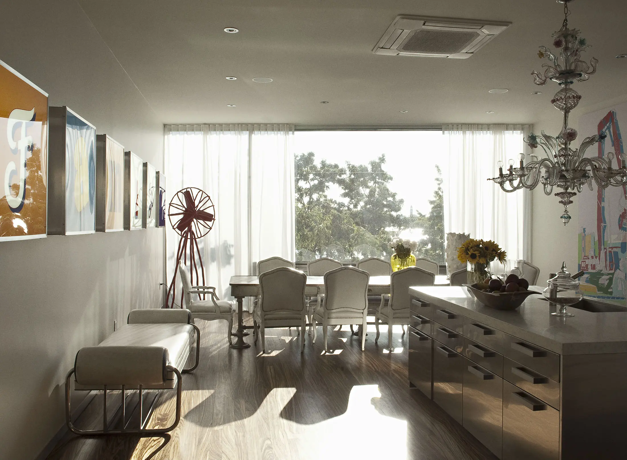 A modern dining room with a wooden floor and large windows letting in natural light. The room features a dining table surrounded by chairs, a metallic kitchen island, and a chandelier. Sunflowers in a vase sit on the island. Artwork and a decorative windmill adorn the walls.