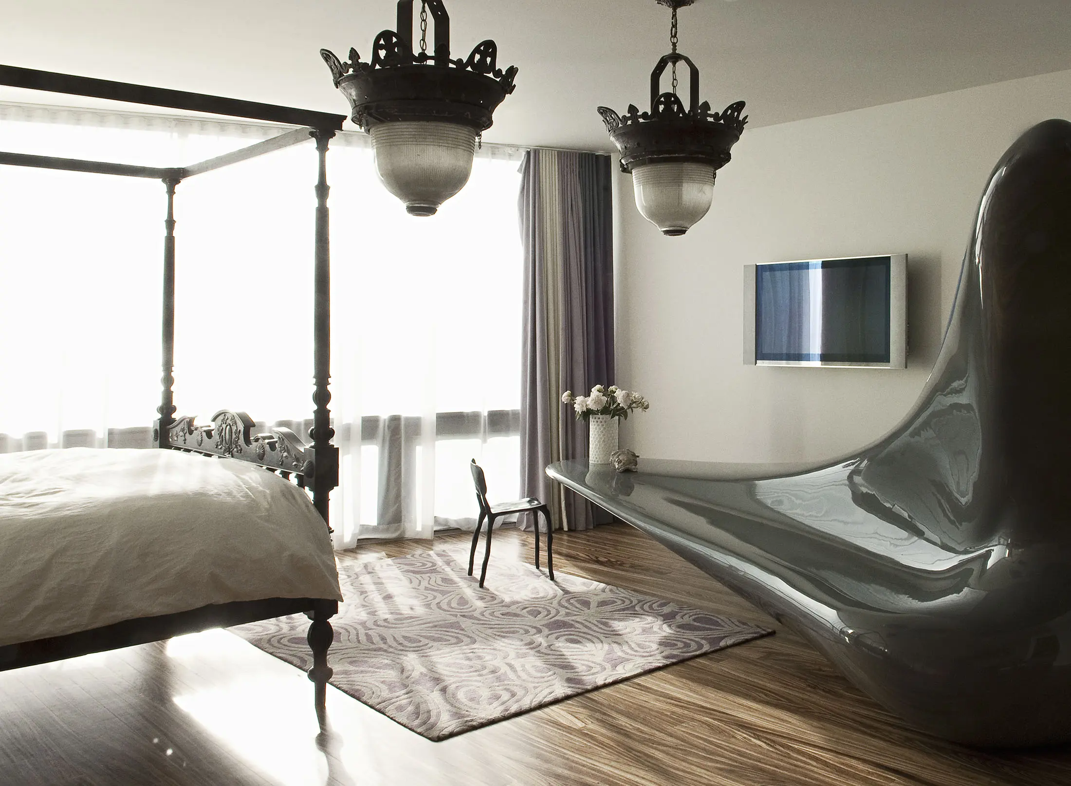 A modern bedroom with a sleek, sculptural gray bathtub. The room features a four-poster bed with white bedding, ornate ceiling lights, a flat-screen TV, and a small black chair next to a vase of white flowers. The floor is wooden with a patterned rug, and large windows with sheer curtains provide natural light.