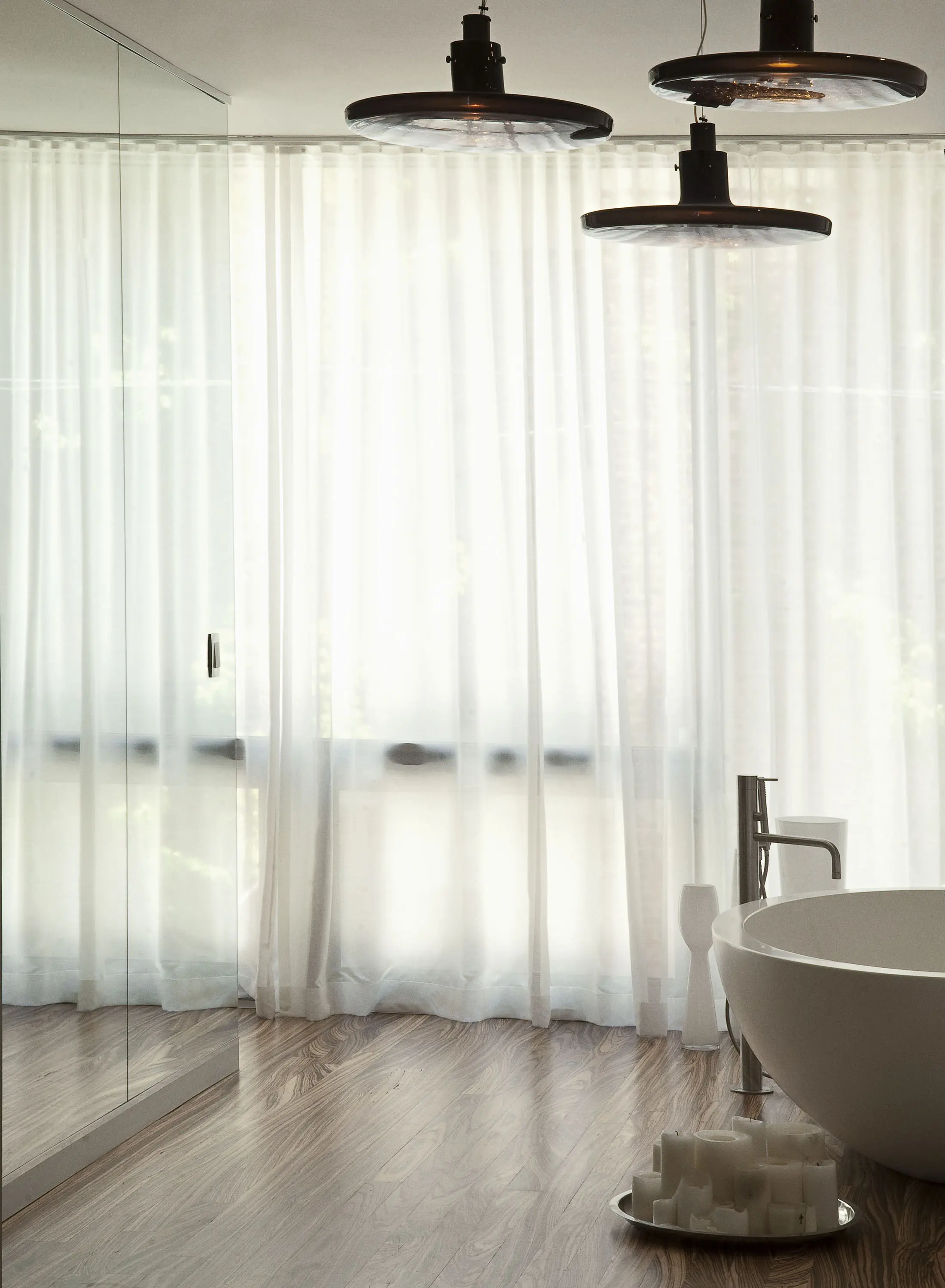 A minimalist bathroom featuring a white freestanding bathtub on wooden flooring. Sheer white curtains cover a large window, allowing soft natural light in. Three modern pendant lights hang from the ceiling. A few decorative candles are placed near the bathtub.