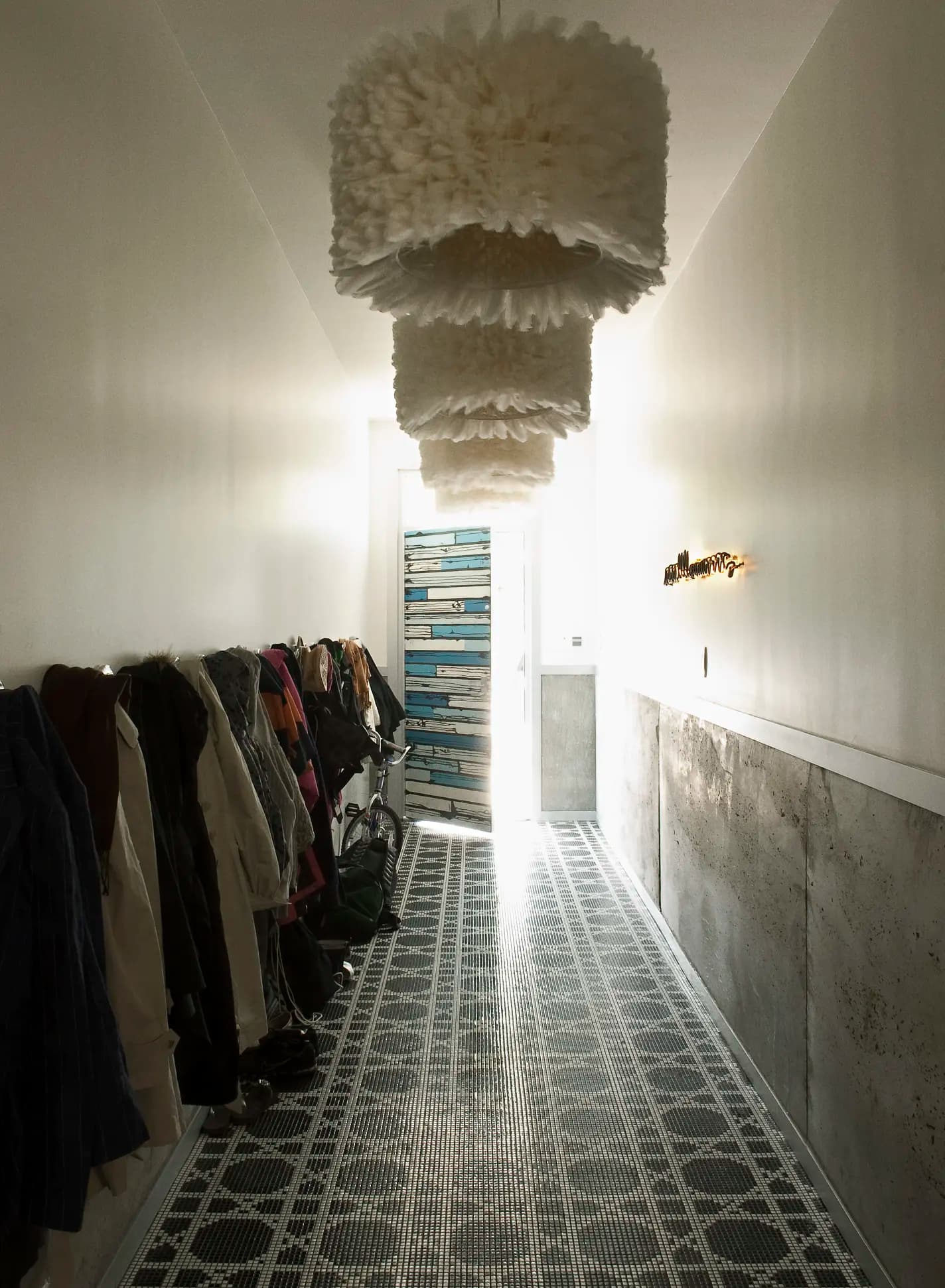 A narrow hallway with a patterned black and white tiled floor leads to a door with stacked picture frames. Coats hang on the left wall; the right wall is bare except for a row of lights. Two large, fluffy white pendant lights hang from the ceiling, casting a soft glow.