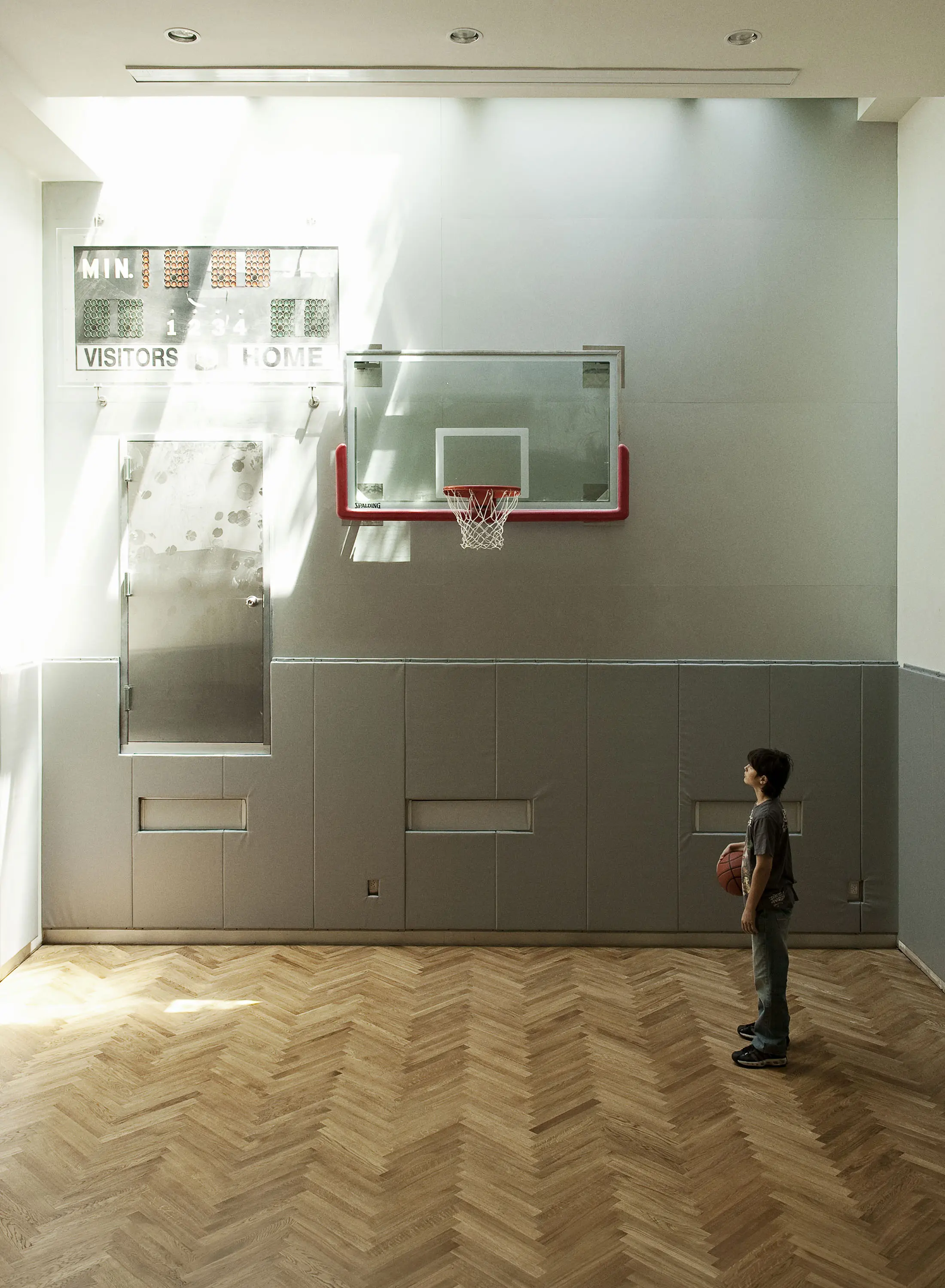 A young boy holding a basketball stands in an indoor court, facing a hoop with a red rim and clear backboard. Sunlight streams in from the left, casting shadows on the parquet floor. A scoreboard is visible above, with the score partially visible. The walls are light-colored.