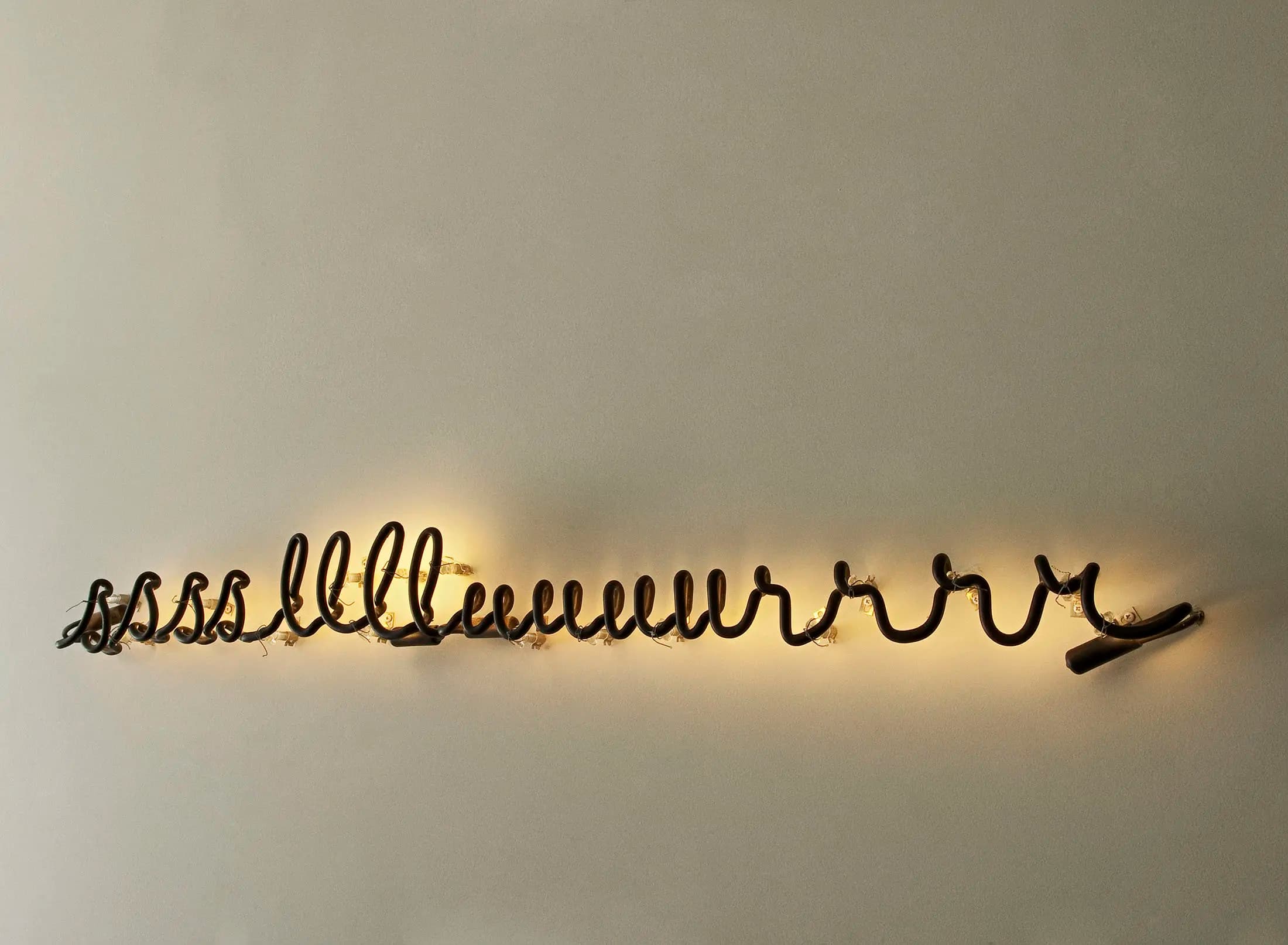 A wall-mounted neon art installation featuring a continuous black wavy line with glowing white light emanating around it. The line varies in height, creating a dynamic, undulating pattern that stands out against the plain beige wall. The artwork creates a warm, ambient glow.