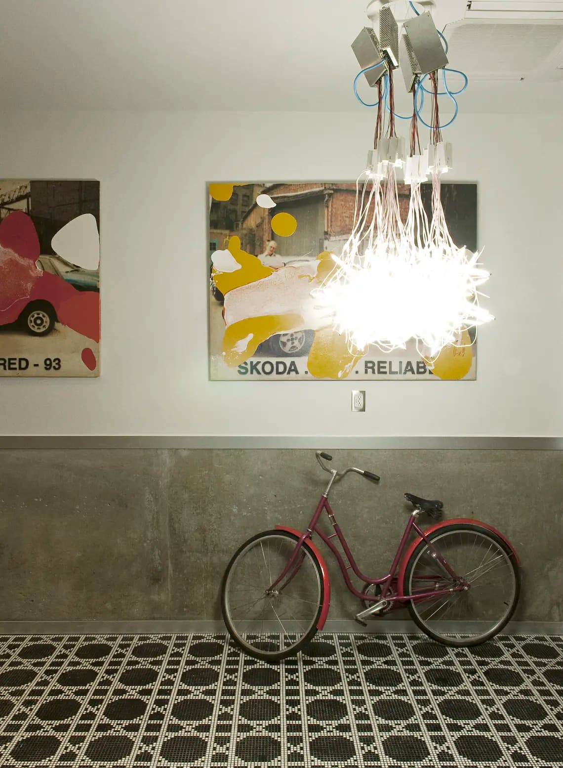 A red bicycle leans against a wall with a concrete lower half and white upper. Above, a bright, modern chandelier hangs, and two colorful abstract paintings with visible text are mounted on the wall. The floor features a black and white patterned design.