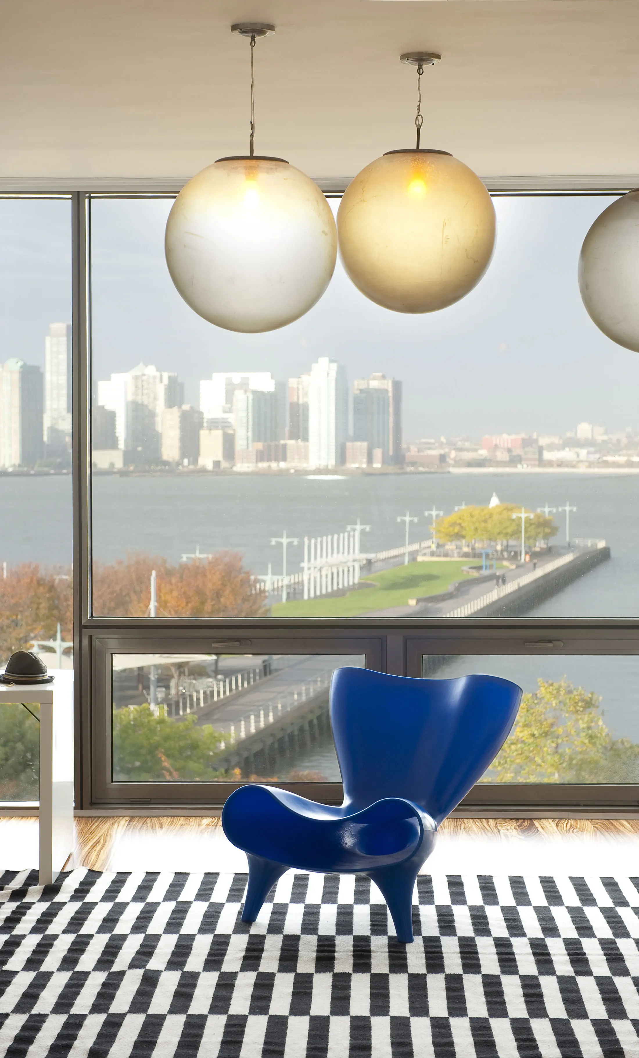 A modern interior featuring a large window with a waterfront cityscape view. A unique blue chair is centered on a black-and-white checkered rug. Above, three spherical pendant lights hang from the ceiling. The scene outside shows buildings and a pier with autumn trees.