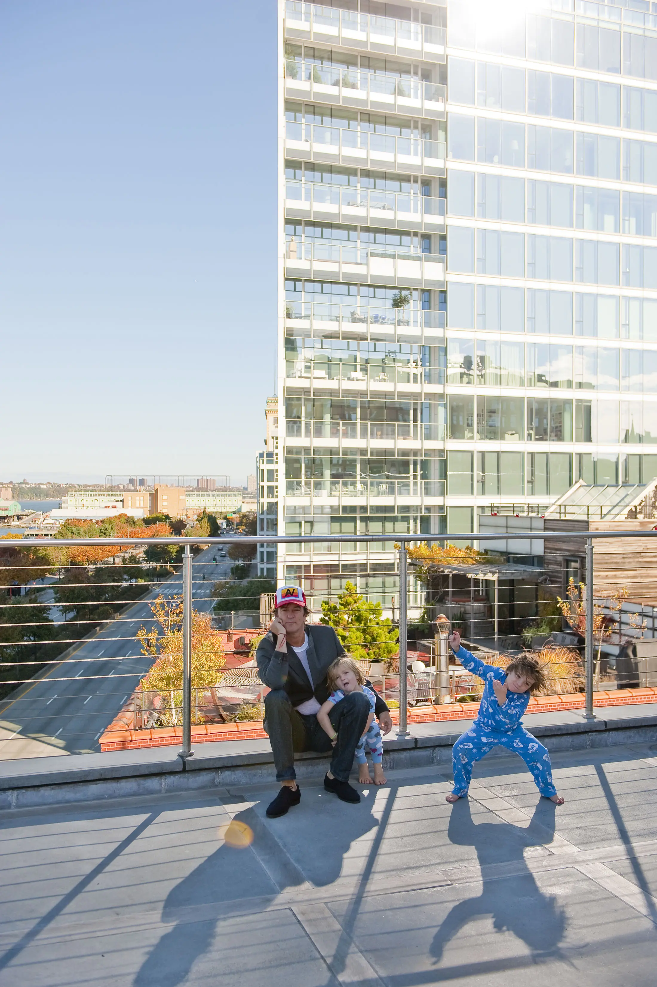 A person crouches on a rooftop terrace, affectionately hugging a child with long blonde hair. Next to them, another child in blue pajamas is playfully posing. The background features a tall, glass-fronted building and clear blue sky. The terrace has metal railings and urban scenery is visible.